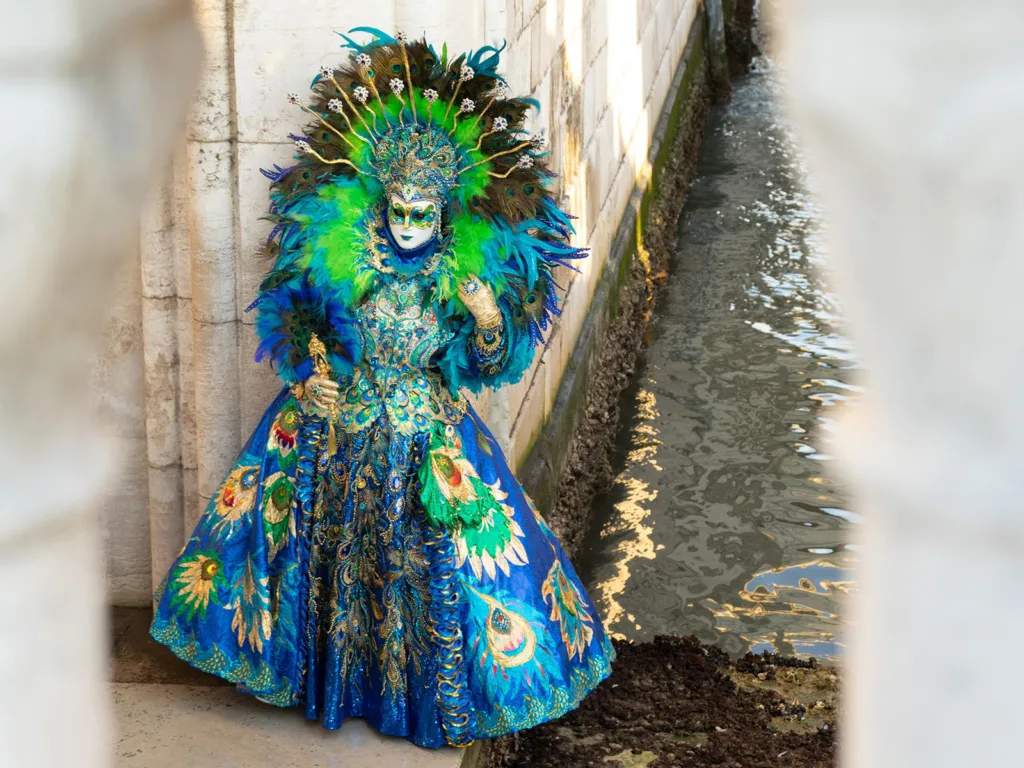 A woman wears a vibrant blue and green dress, mask, and elaborate headdress in Venice, another top reason to travel to Italy | Davidsbeenhere