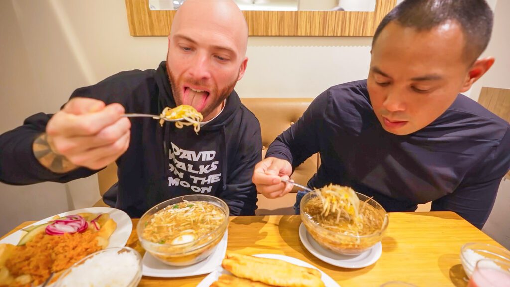 David Hoffmann and his cameraman Mike take their first bites of saoto soup, a Suriname food containing chicken, noodles, and bean sprouts | Davidsbeenhere