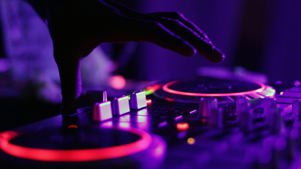 A closeup of a DJ's deck, with several buttons, sliders, and glowing turntables | Davidsbeenhere